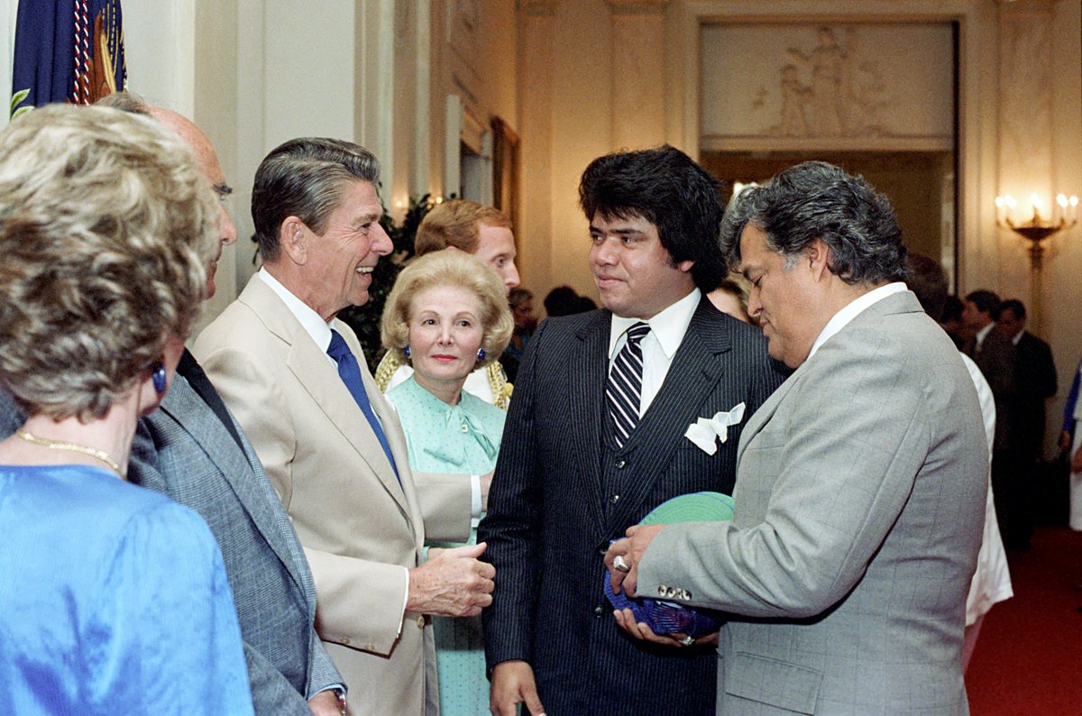 Color photo of President Ronald Reagan in a suit and tie tackled to a  man dressed in a pinstriped suit and tie with a pocket square. They are surrounded by people.
