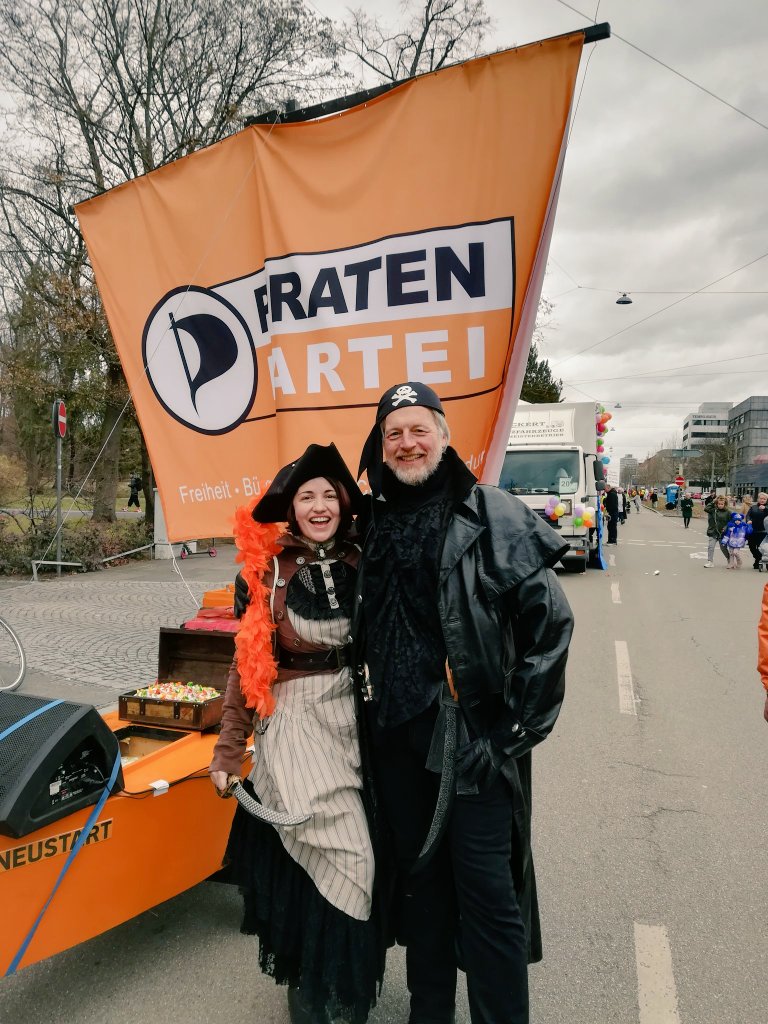 Zwei Personen sind als Piraten verkleidet Anja Mitte Rock Dreispitz Säbel und Orangen Federn und ein männlicher Pirat mit Gehrock im Hintergrund ein Boot mit Rädern und einer großen Orangen Segel mit der Aufschrift Piratenpartei