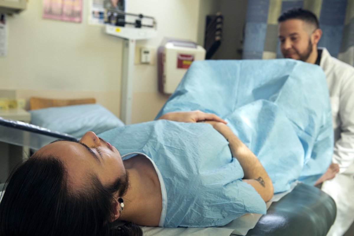[ID: a patient with long brown hair and earrings wears a hospital gown and lies on a doctors' table. A doctor sits near their feet, wearing a white lab coat.]