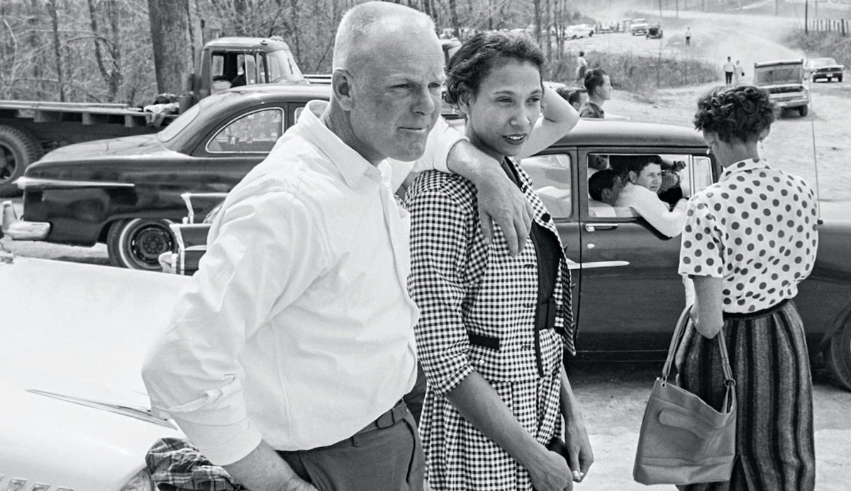 An old photograph of Richard and Mildred Loving. They are standing up outside, and Richard has his arm on Mildred's shoulder.