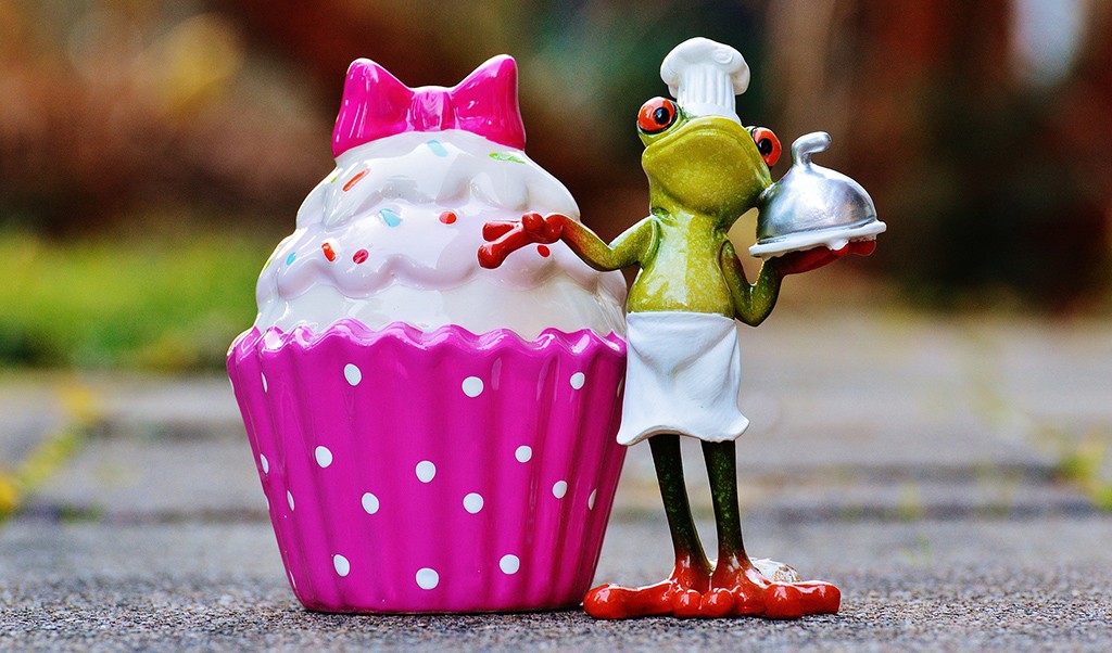 a frog figurine standing next to a giant cupcake holding a platter. wearing a chef's hat and apron