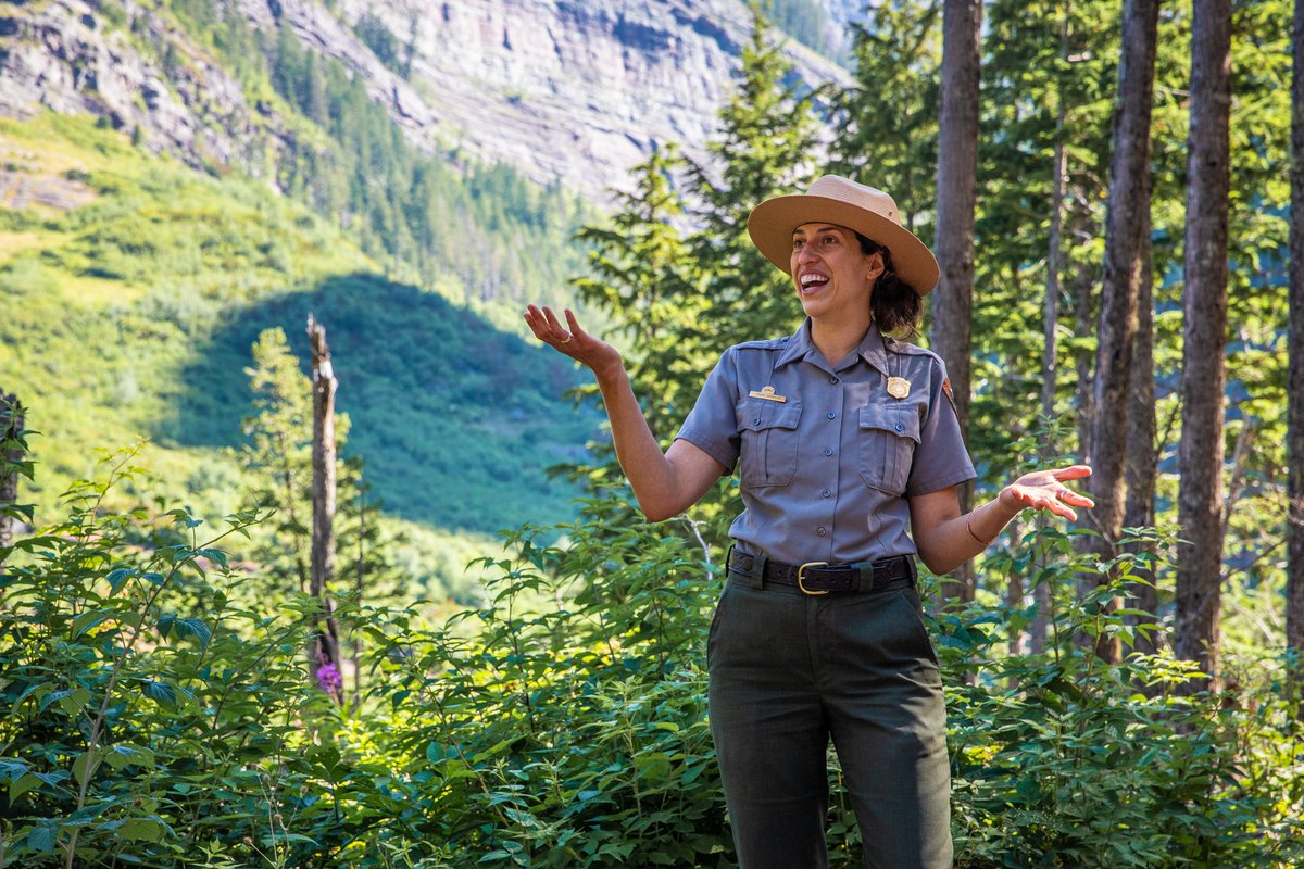 A national park ranger smiles and shrugs their shoulders in a forest. https...