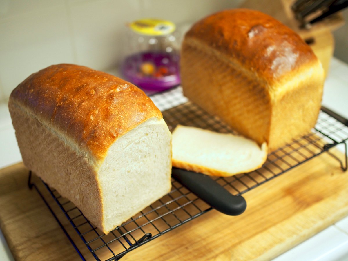 Two loaves of sourdough sandwich bread on a cooling rack. One is sliced to reveal a creamy white crumb. The tops are mottled, golden brown, and buttery. They look good, is what I'm trying to say. They're really good.