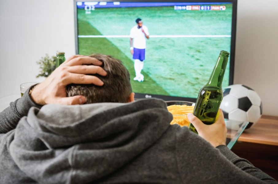 This is the closest-to-reality stock photo of a football fan I could find. In utter despair. Wearing a hoodie. Multiple drinks in front of him. Has already smashed a full bowl of chips and has another ready. But no real football fan would place a ball in front of the screen.