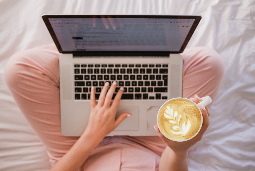 a woman sitting on a bed holding a cup of coffee and typing on a laptop with the other hand
