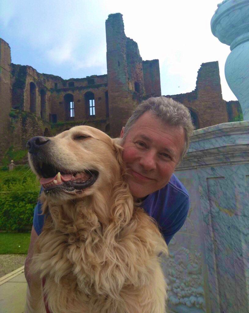 Pip is with Family Dad in the grounds of Kenilworth Castle. Dad is snuggling up behind Pip  in front of the castle walls. Pip is looking to the left with a smile on his face, while Dad is in a blue t-shirt and crouching down with a smile on his face.
