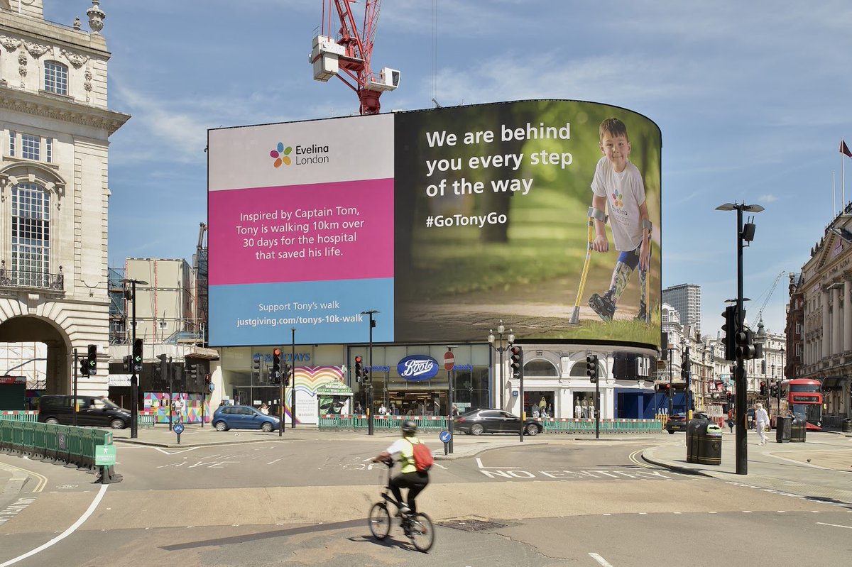 A  huge sign in Piccadilly Circus with the Evelina London logo and a picture of Tony walking. Words on the sign say "We are behind you every step of the way #GoTonyGo".