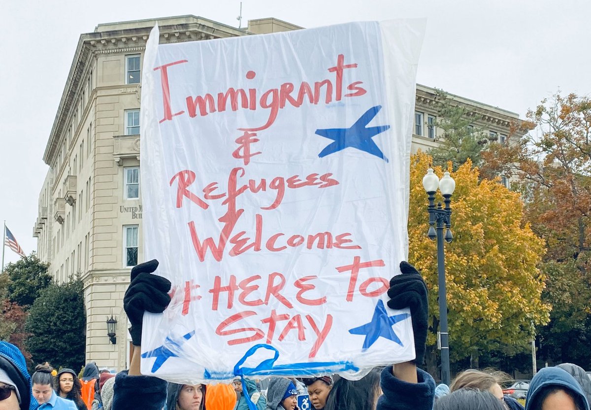 Protest sign outside the Supreme Court says "Immigrants and refugees welcome. #HereToStay."