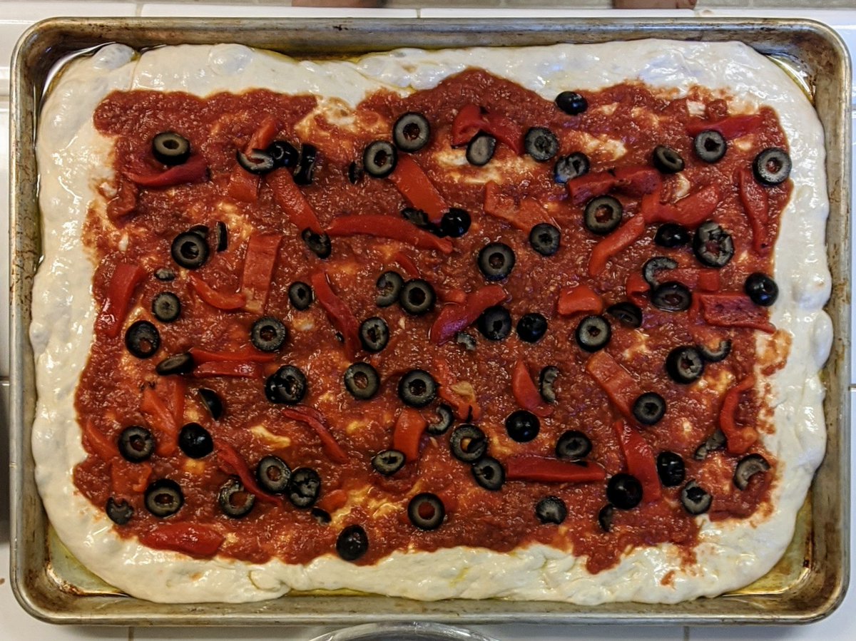 Raw pizza dough, topped with sliced olives and roasted red peppers. Oil pools in the corner of the steel sheet pan.