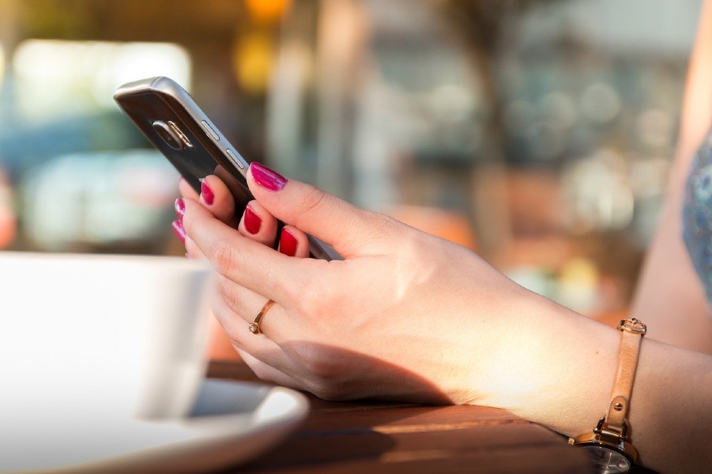 a woman hlding a smartphone sitting at a table with a cup of coffee by her side
