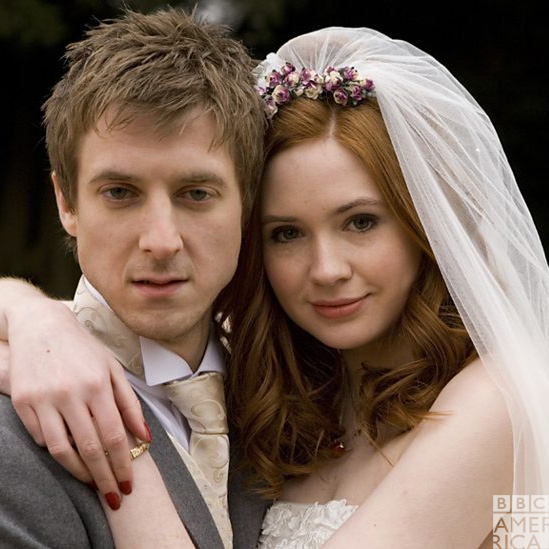 Rory and Amy looking at the camera, smiling. Rory's wearing a gray suit, Amy's wearing a white dress and veil.