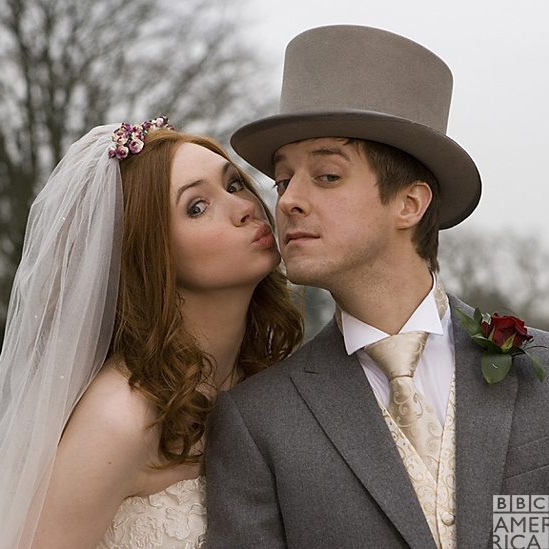 Amy is leaning in to kiss Rory's cheek. She's wearing a white wedding dress and veil, Rory's wearing a gray suit and top hat.