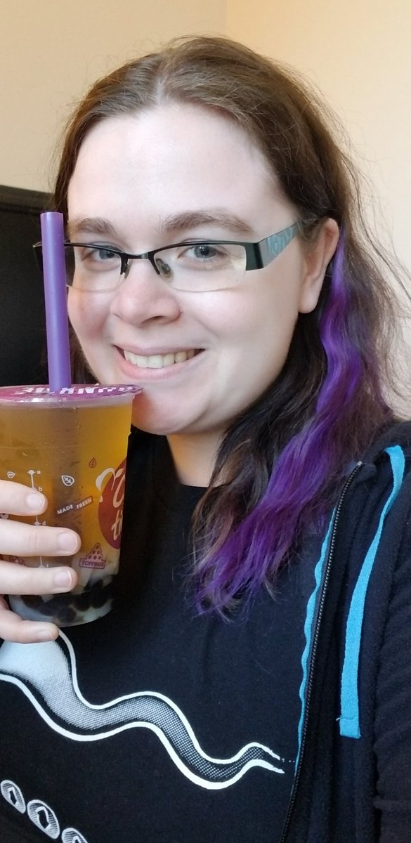 White girl with brown and purple hair smiles at the camera holding a bubble tea next to her face.