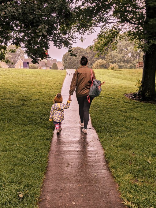 Mother and daughter walk hand in hand along a path leading to a duck pound whilst green trees frame the picture.