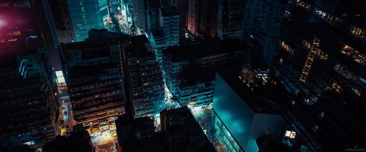Photography by Liam Wong of Hong Kong at night. Looking down over the city from above. We see older high rise residential buildings surrounded by modern architecture. It feels reminiscent of establishing shots in sci-fi movies. It is teal and yellow in color and has a pink highlight from a light.