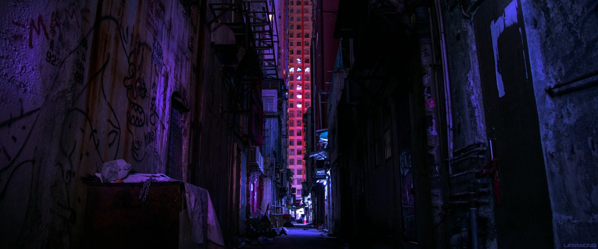 Photography by Liam Wong of Hong Kong at night. An old backalley covered in graffiti. It is well lit with a spotlight on the end of the alley. There is a large building in the distance which covers the sky. The lights have been altered into the shape of a cross.