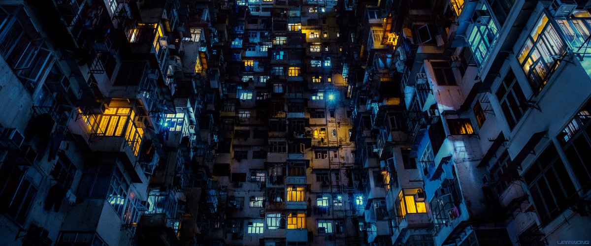 Photography by Liam Wong of Hong Kong at night. The famous Mansions - old style residential building. The camera is looking upwards and we feel the scale of the building, which is made up of many floors and lights with caged windows. It is blue and yellow in color.