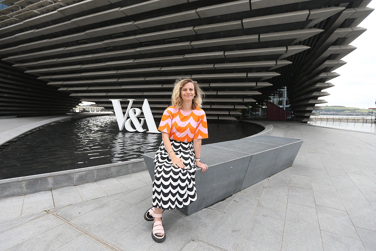A woman wearing an beautiful orange and white striped shirt with white and black striped skirt sitting on the concrete bench outside of V&A Dundee.