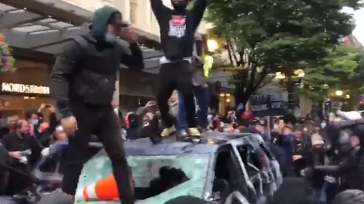 With a hard cut, the video switches to the busted out roof of a car. Protesters dance atop it. Later, it is seen on fire.