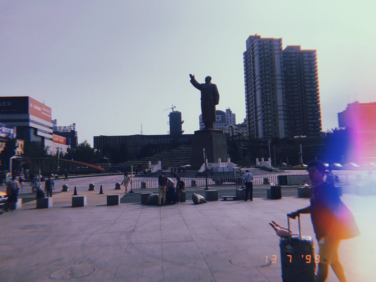 Photo taken outside of Dandong train station where a large statue of Mao, with his right hand raised and head poised to the skies, towers above all the local shops.