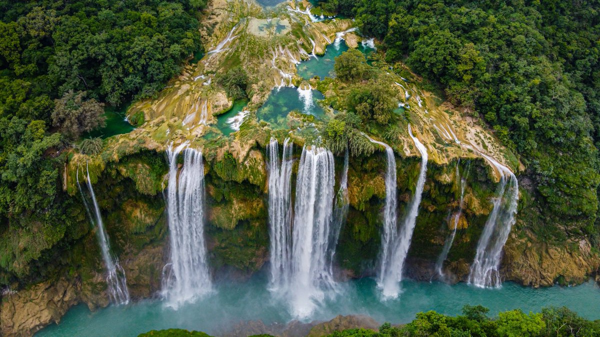 An aerial view of a teal-blue waterfall emerging from a lush green forest.