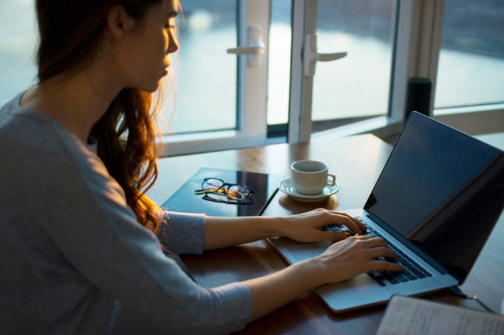 a woman sitting at her desk looking down at a laptop with a cup of coffee by her side
