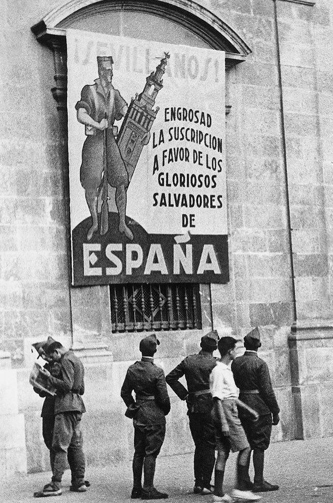 Nationalist soldiers in Seville during the Spanish Civil War looking up at a fundraising poster. The poster shows a soldier posing with the city’s iconic giralda belltower, with the text reading – “Sevillanos! Increase your subscription for the glorious saviours of Spain”.⁣