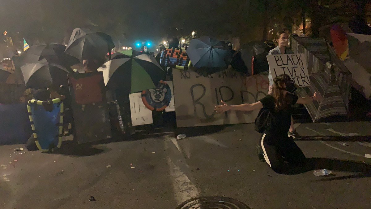 In front of the line of DIY shields and umbrellas, two young people stand unarmed without helmets, gas masks, or even long sleeves/pants.