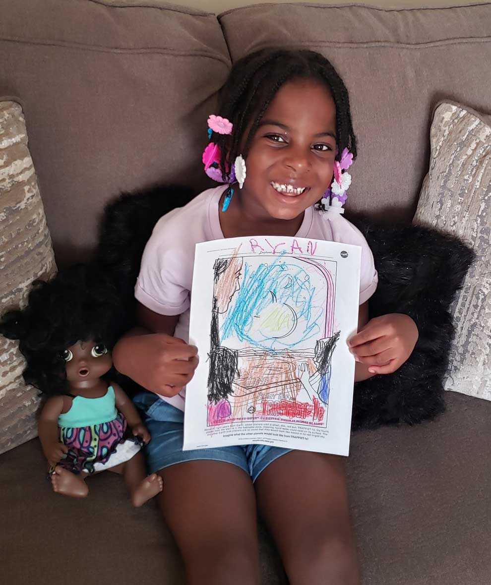 An adorable little girl holds her coloring page for the TRAPPIST-1e exoplanet poster. She has a little doll next to her.