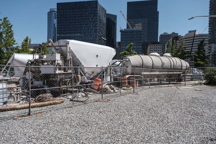 Concrete pumping trucks staged in a gravel yard protected by fencing, with Seattle skyline in the background