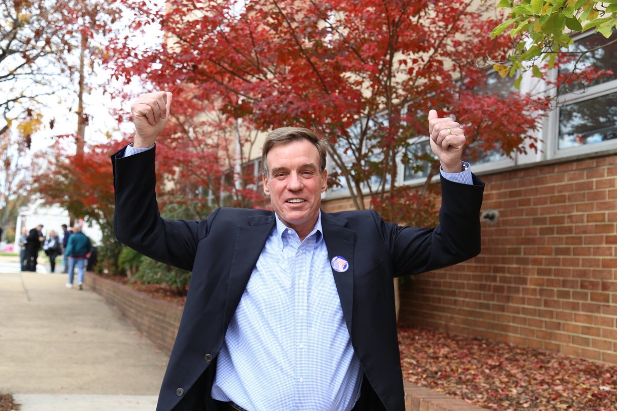 Senator Mark Warner, who is wearing a sticker indicated he voted, stands outside of his polling location on Election Day in 2014. He is raising his arms and giving a thumbs up.