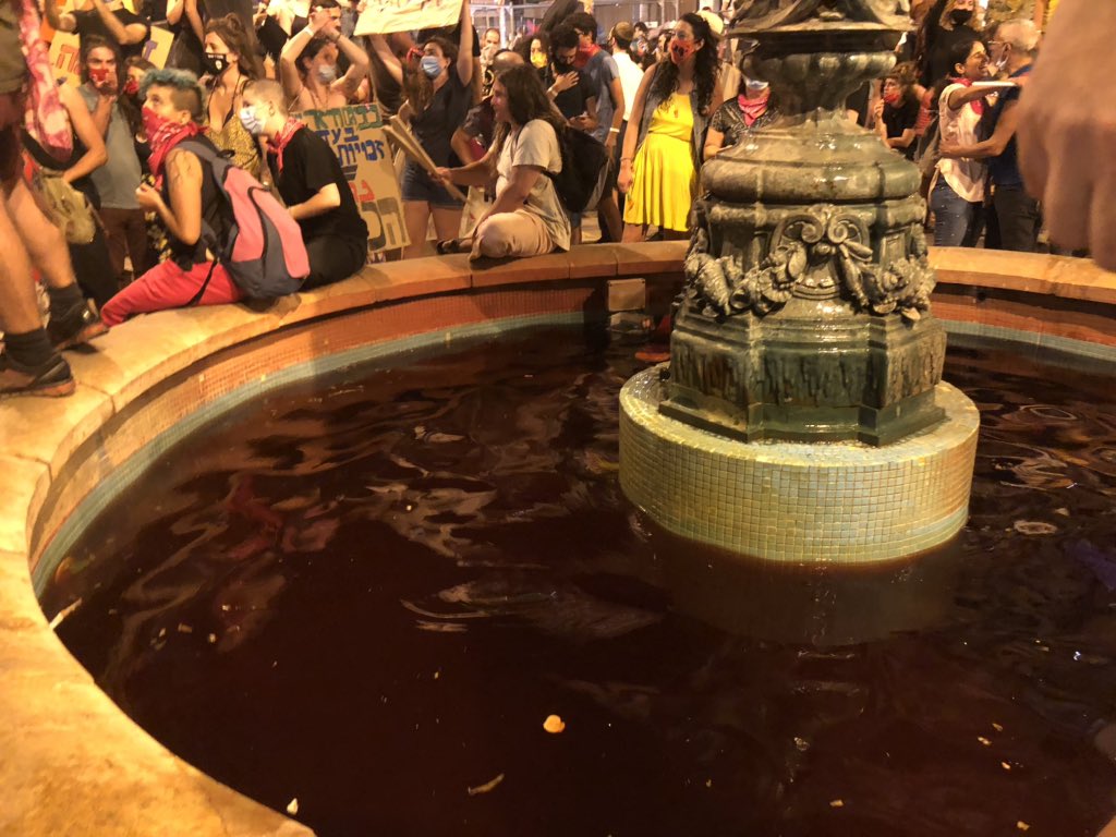 A pride flag flies next to an Israeli flag and several black flags above the fountain in the centre of the square. Meanwhile, the water in the fountain had been dyed red