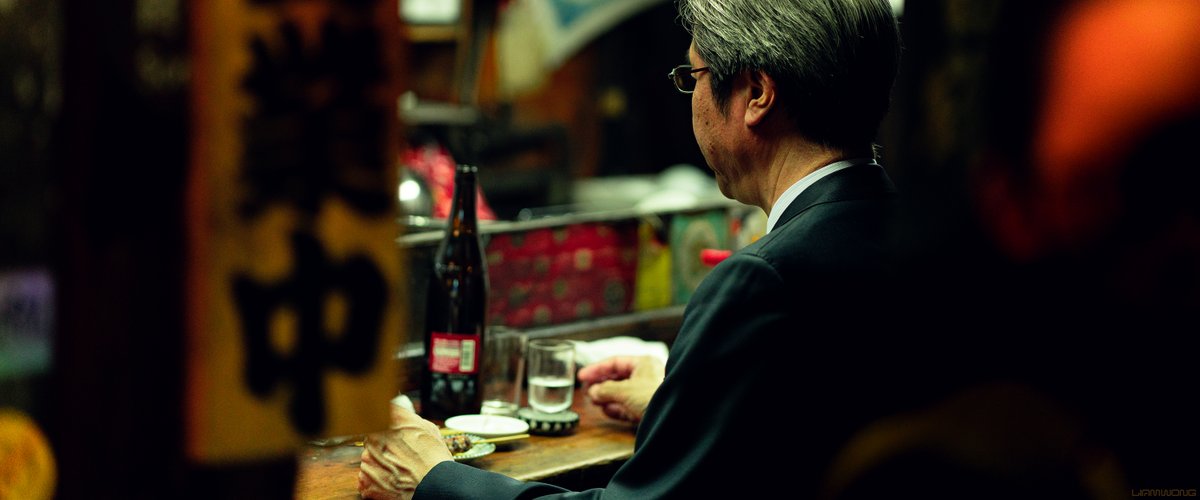Photography by Liam Wong of Tokyo at night. A salary man is sat at a table with a bottle of sake and is eating yakitori. He is seen from behind.
