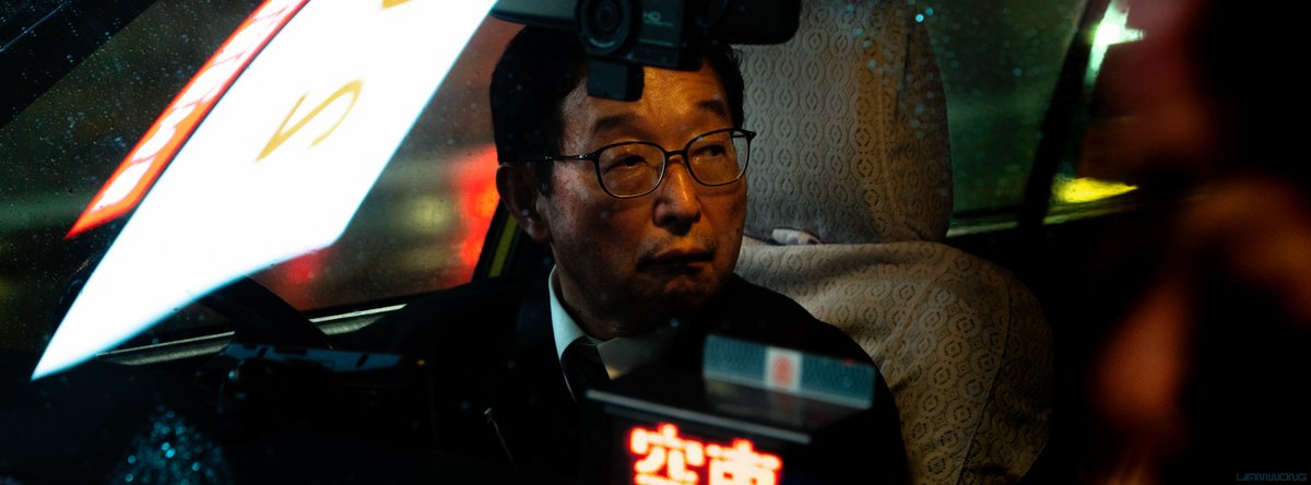 Photography by Liam Wong of Tokyo at night. The interior of a taxi driver - captured from the outside as he glances to his left at Shibuya crossing.