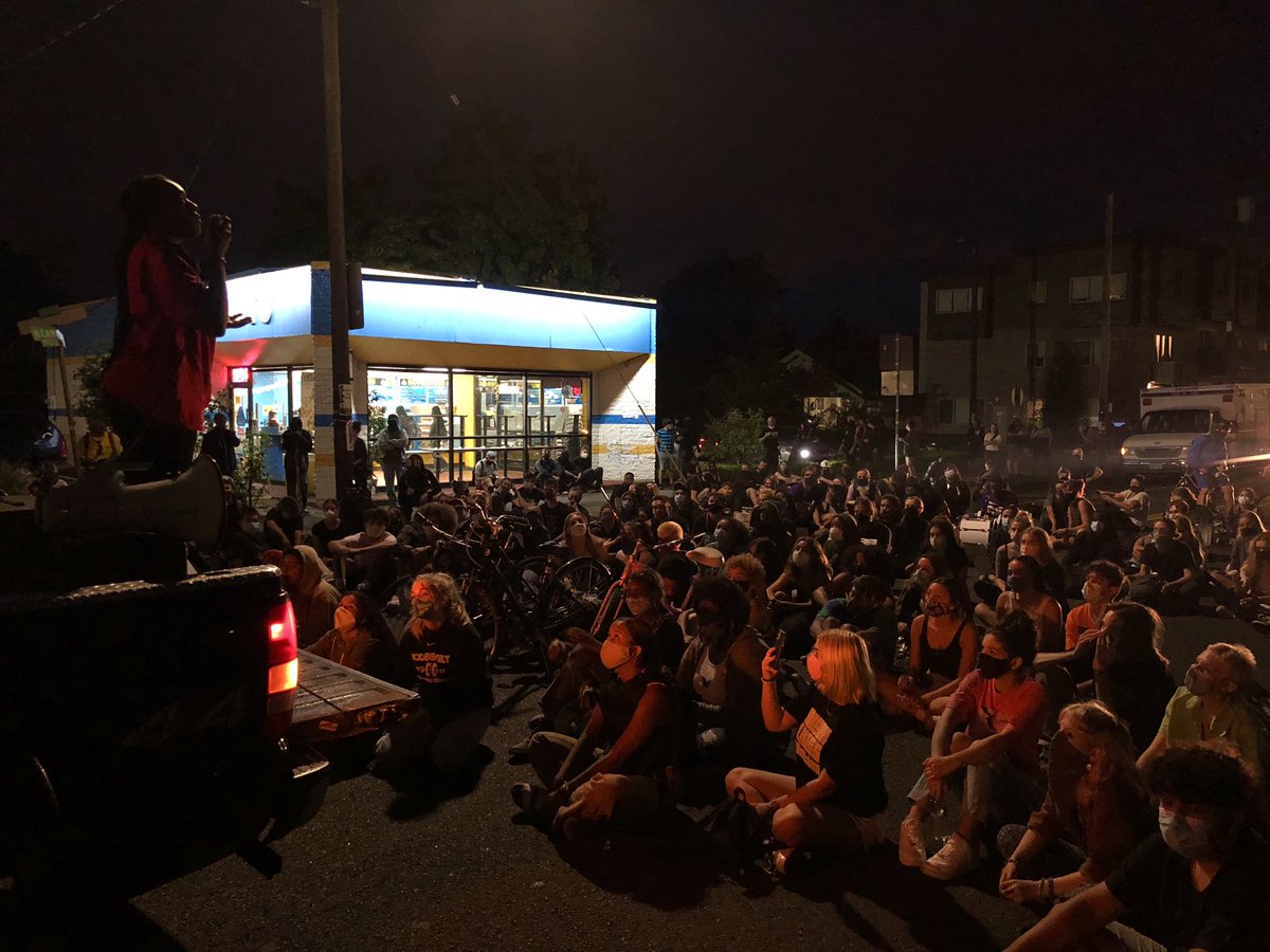 Vehicles are parked on N. Denver and Elmore avenues blocking traffic onto the stretch of Lombard in front of the Portland Police Association building. The crowd is sitting and listening as PDXBYM leaders take turns speaking into a megaphone.