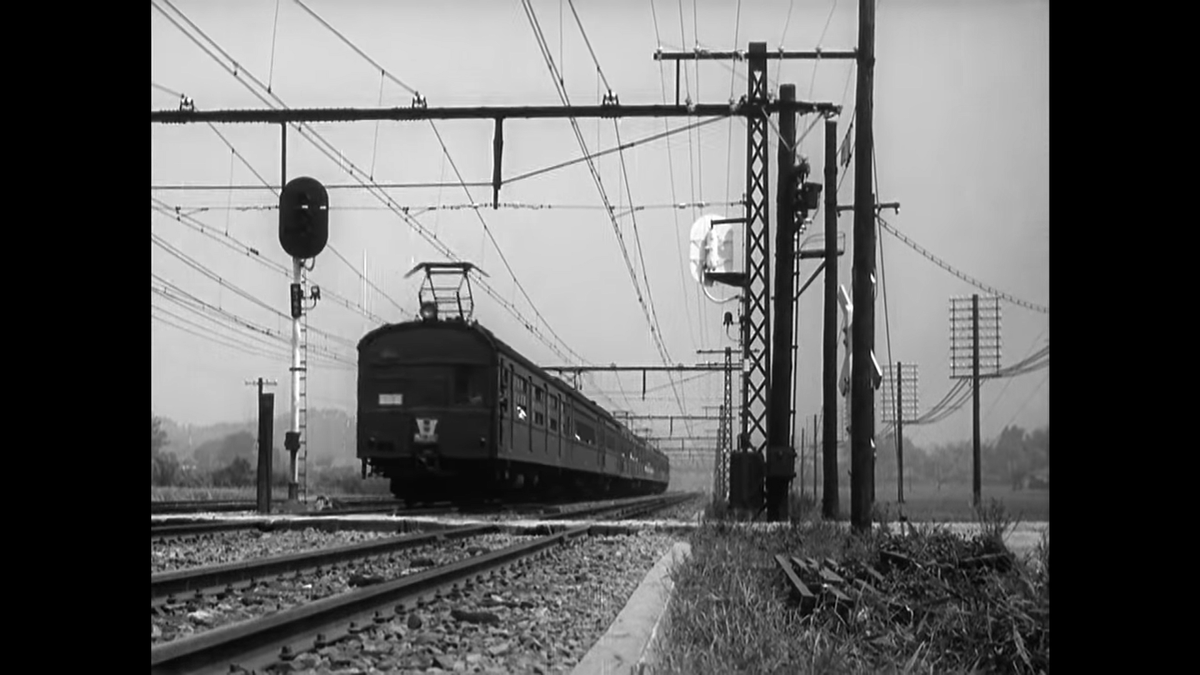 This train shot is characteristically long (relatively speaking) at 15 seconds. We see the whole train go by, but Ozu also holds on the shot until the train is practically out of sight. Note, too, everything going on in this simple train shot! Even some wood in the grass.