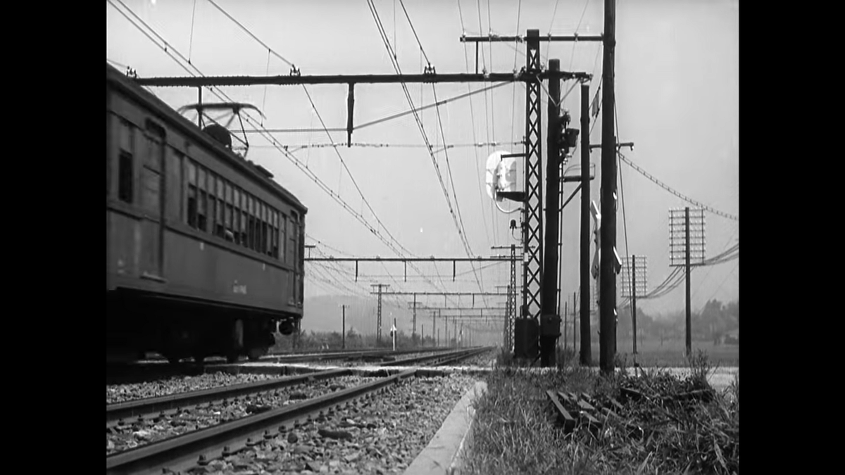 This train shot is characteristically long (relatively speaking) at 15 seconds. We see the whole train go by, but Ozu also holds on the shot until the train is practically out of sight. Note, too, everything going on in this simple train shot! Even some wood in the grass.