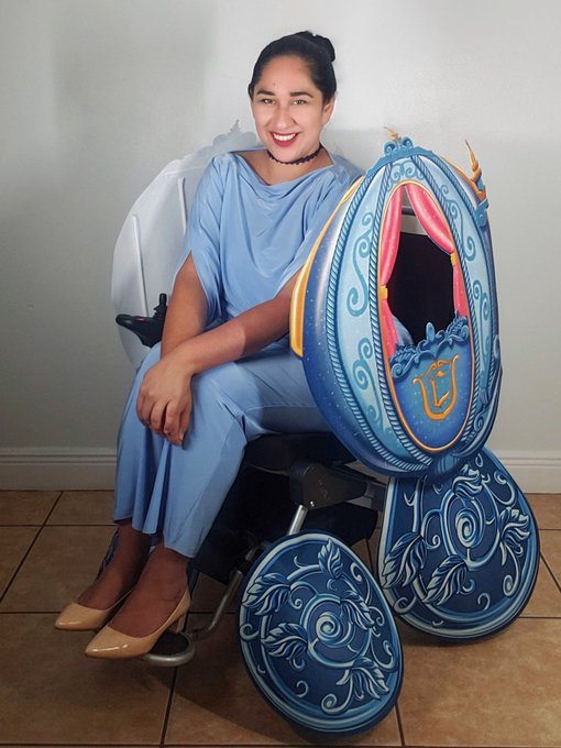 Annie is wearing a light blue jumpsuit, black choker necklace, and her hair in a high bun. She is sitting in her wheelchair with a wheelchair cover that is tied to the sides and hanging over the tires of her wheelchair in order to look like Cinderella’s Coach.