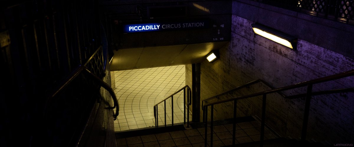 Photography by Liam Wong of subways at night. This image is of Picadilly Circus in London. A yellow light from the halogens light up the stairs. Purple lights from the neons surrounding compliment the color.