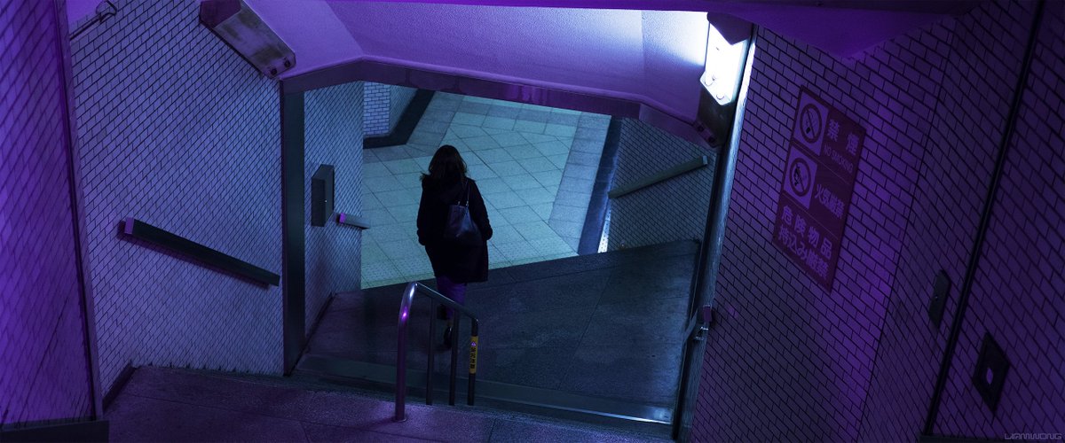 Photography by Liam Wong of subways at night. This image is of Shinjuku in Tokyo. A purple flash lights up the interior as a woman walks down the stairs to the station.