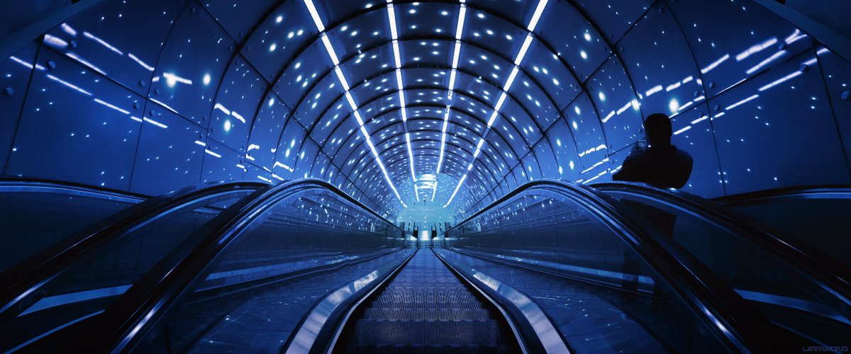 Photography by Liam Wong of subways at night. This image is of Warsaw. Futuristic looking lights shot looking downwards. It feels like a hypervision tunnel / style image. A man is on his phone as he moves down the escalators.