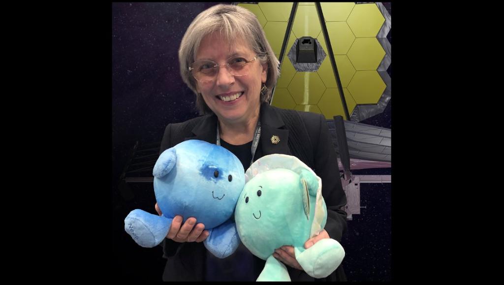 Dr. Heidi Hammel poses in front of a James Webb Space Telescope background, holding plush toys of Uranus and Neptune.
