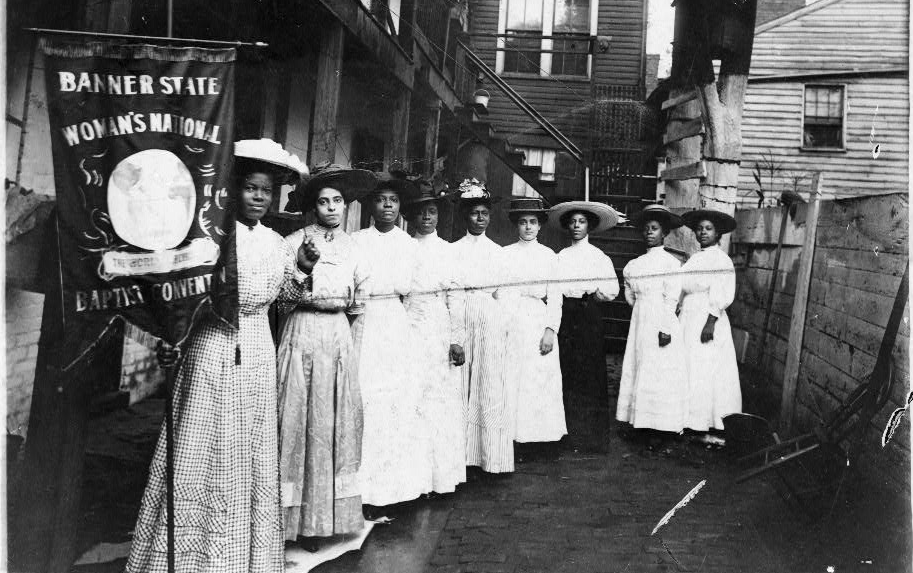 Black and white photo: Suffragists pose, carrying a banner. 