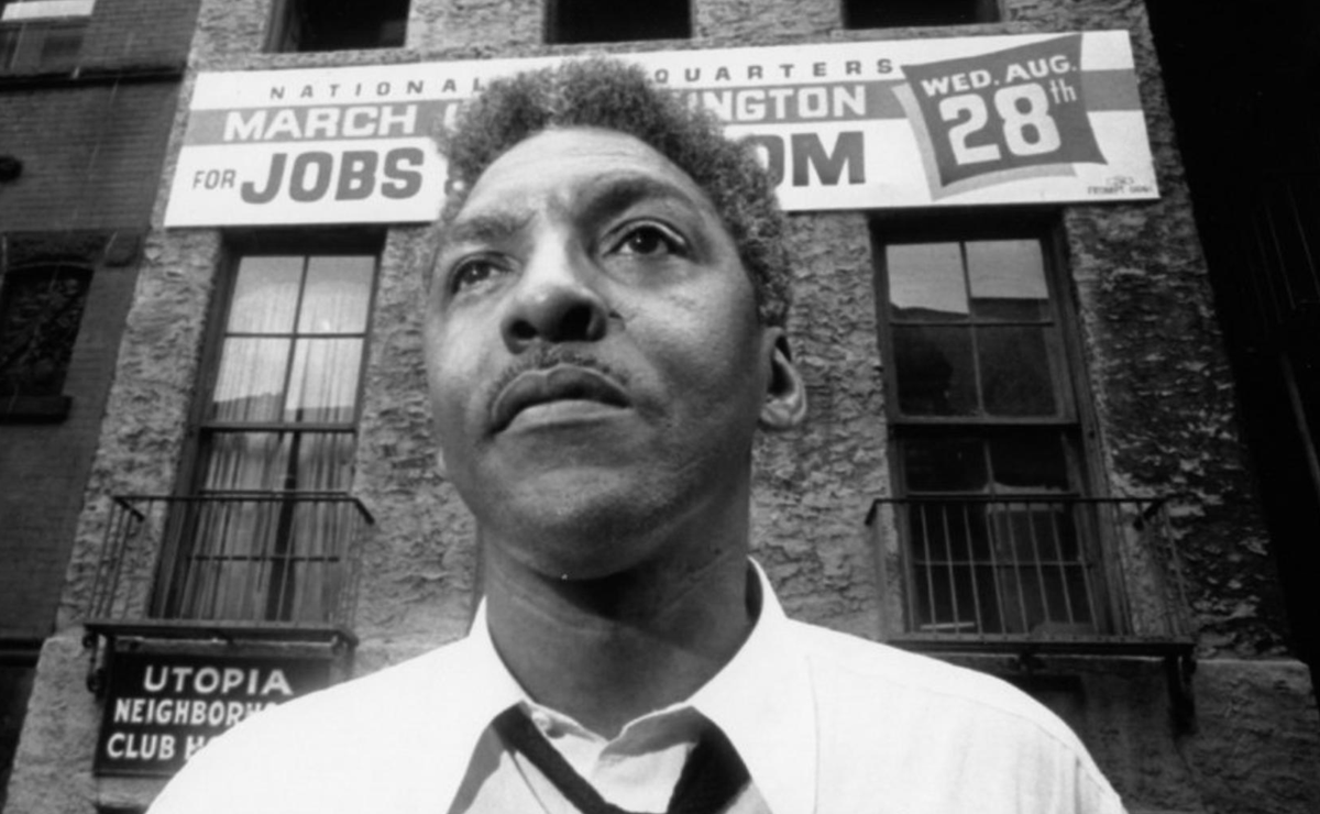 Photo of Bayard Rustin, who is standing in front of a banner promoting the 1963 March on Washington for Jobs and Freedom.