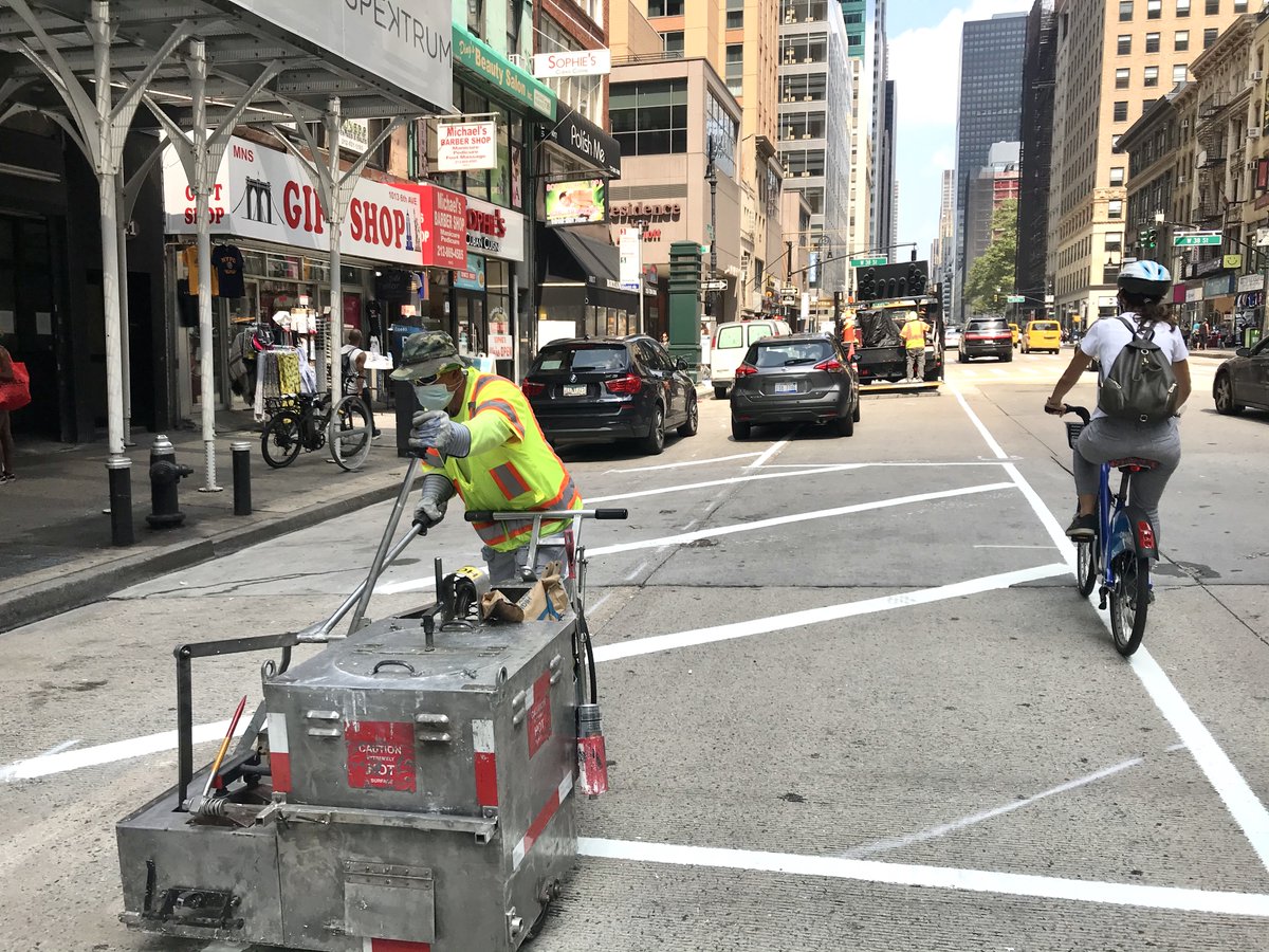 A person in yellow and orange safety gear and a mask operates a machine to paint white lines on a roadway. A person wearing a helmet passes by on a citi bike. 