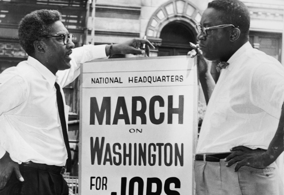 An image of Bayard Rustin standing next to a sign that says "National headquarters, March on Washington for Jobs and Freedom."