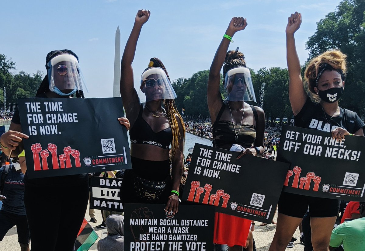 Four marchers at the 2020 March on Washington raise their fists and hold up signs that say "the time for change is now," "maintain social distance, wear a mask, use hand sanitizer, protect the vote," and "get your knee off our necks!"