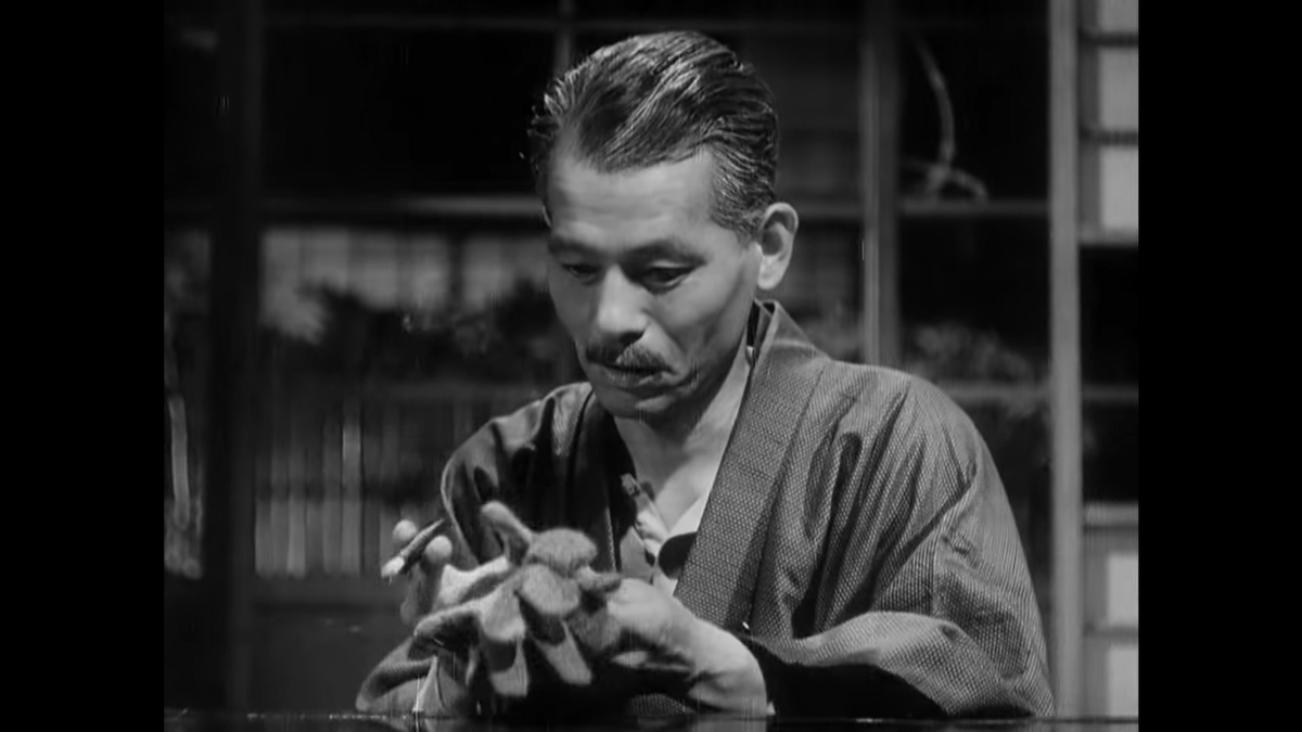 Noriko has brought Shukichi his gloves, which he had misplaced. This guy would lose his head if Noriko didn’t screw it on for him. This shot is head on across the table from Shukichi, but in between the POV of the two other characters.