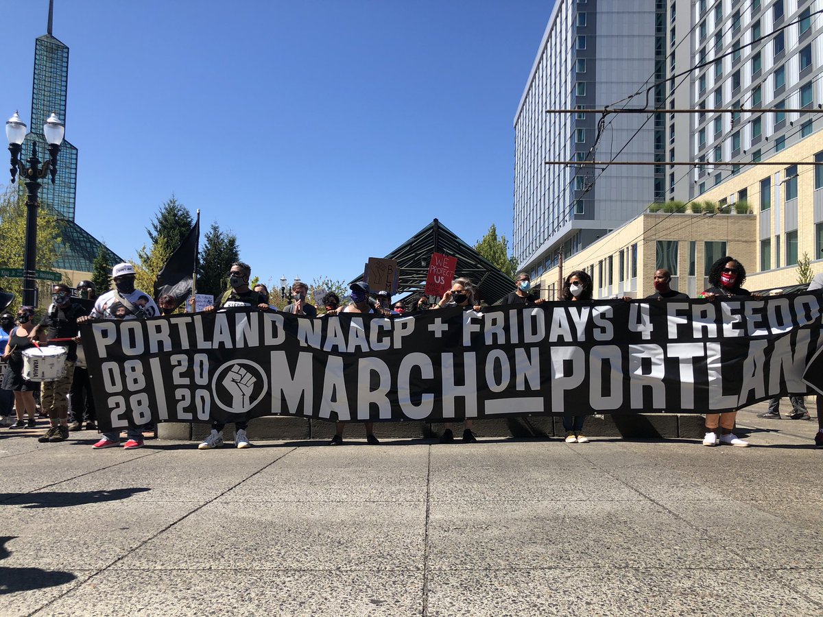 The crowd has gathered on NE MLK Jr. Blvd. and Holladay St. and is preparing to march. A group at the front holds a black banner reading “PORTLAND NAACP + FRIDAYS 4 FREEDOM / 08-28-2020 / MARCH ON PORTLAND”