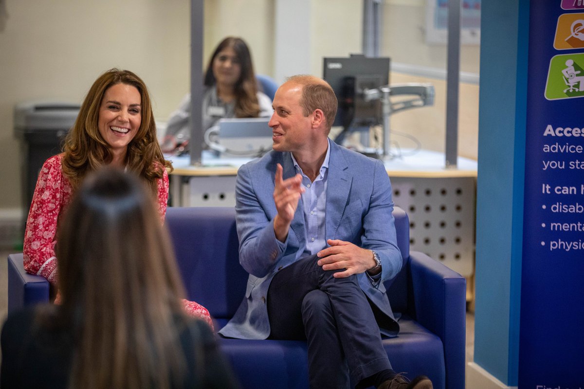 HRH The Duke and Duchess of Cambridge sit and chat with staff inside the jobcentre. They are facing the camera and laughing.